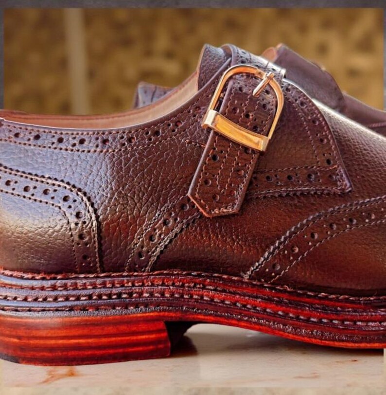 K&ouml;nnte beinhalten: Nahaufnahme eines braunen Lederschuhs mit dekorativer Schnalle. Der Schuh hat aufwendige N&auml;hte und Perforationen, mit einer rotbraunen Sohle und einem Absatz. Die goldfarbene Schnalle verleiht dem Schuhwerk einen Hauch von Eleganz.