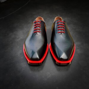 May include: A pair of black leather shoes with red soles and red laces. The shoes have a classic design with a pointed toe and a low heel. The shoes are displayed on a dark surface.