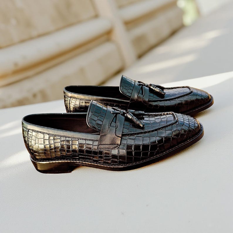 May include: A pair of black leather loafers with a crocodile skin texture. Each shoe features a decorative strap with a tassel. The shoes are displayed on a white surface, showcasing their glossy finish and classic design.