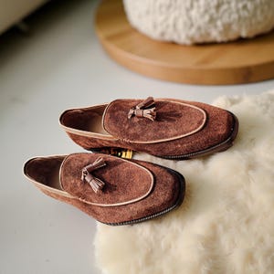 May include: A pair of brown suede loafers with a tassel detail. The shoes have a rounded toe and a light brown trim. The loafers are resting on a white, fluffy surface.