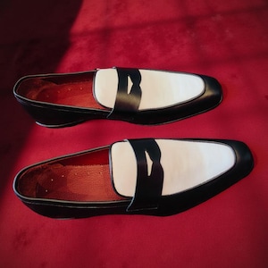 May include: A pair of black and white leather loafers. The shoes feature a white vamp, black trim, and a black strap. The interior is red, and they are set against a red background. A classic footwear design.