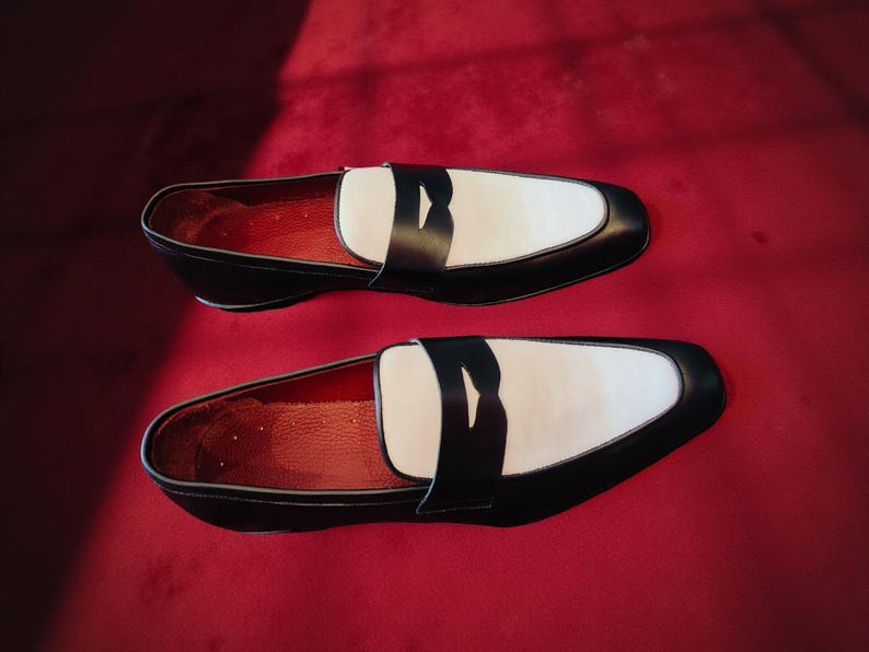 May include: A pair of black and white leather loafers. The shoes feature a white vamp, black trim, and a black strap. The interior is red, and they are set against a red background. A classic footwear design.