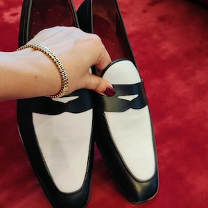 May include: A pair of black and white leather loafers. The shoes have a black body with white vamps and a black band across the top. The interior is red. The shoes are on a red surface.