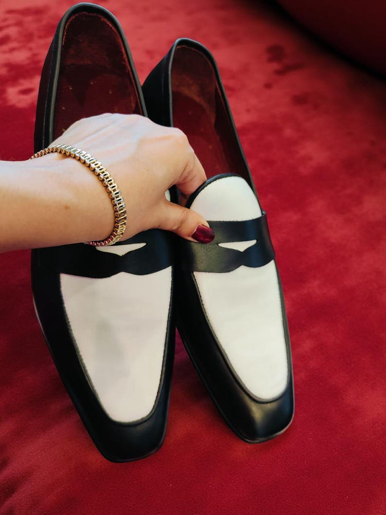 May include: A pair of black and white leather loafers. The shoes have a black body with white vamps and a black band across the top. The interior is red. The shoes are on a red surface.