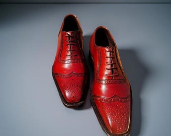 May include: A pair of red leather dress shoes with brogue detailing. The shoes have a classic Oxford style with a lace-up closure and a polished finish. The shoes are displayed on a solid blue background.