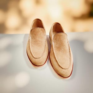 May include: A pair of tan suede loafers with white stitching and a red-brown sole. The shoes are displayed on a white surface, with a blurred background of warm tones.