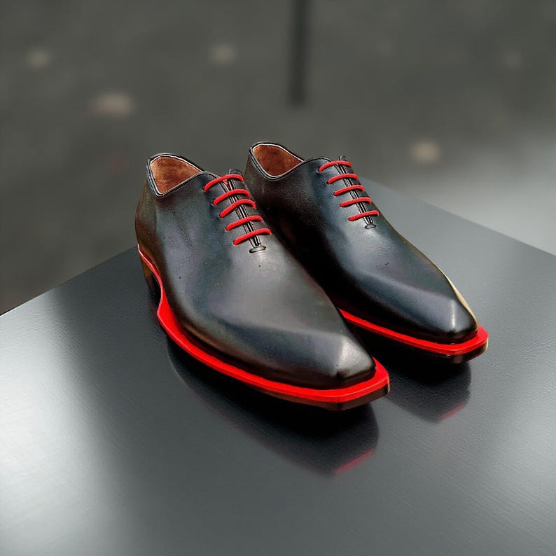 May include: A pair of black leather dress shoes with bright red laces and a red sole trim. The shoes are displayed on a dark surface, showcasing their sleek design and contrasting colour accents. The shoes have a pointed toe.