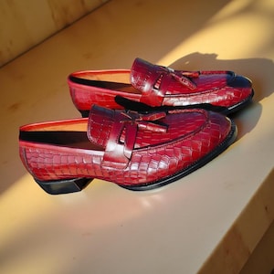 May include: A pair of burgundy leather loafers with a crocodile texture. The shoes have a classic design with a tassel detail and a black sole. The loafers are displayed on a light-colored surface.