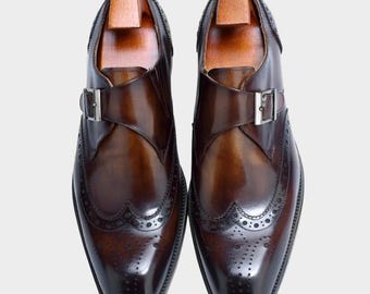 May include: A pair of brown leather dress shoes with a monk strap buckle closure. The shoes feature brogue detailing along the edges and a wooden shoe tree inside. The leather has a polished, glossy finish.