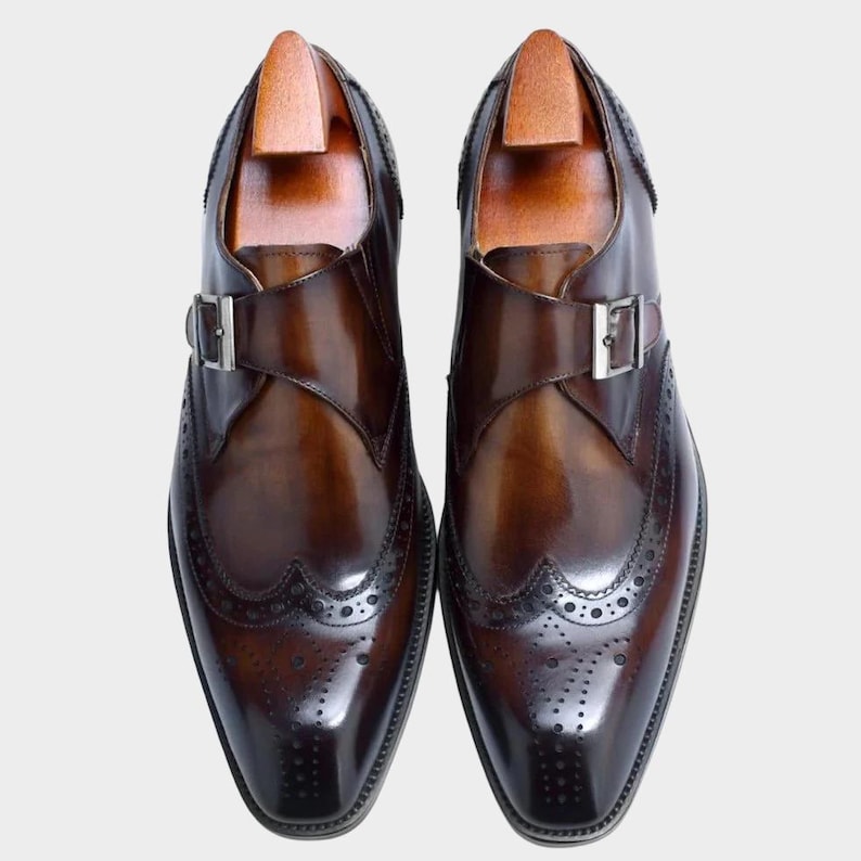 May include: A pair of brown leather dress shoes with a monk strap buckle closure. The shoes feature brogue detailing along the edges and a wooden shoe tree inside. The leather has a polished, glossy finish.