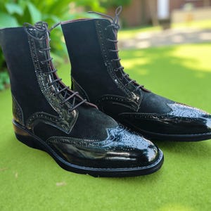 May include: A pair of black leather and suede lace-up boots. The boots have a glossy toe cap and heel, with a suede upper and decorative perforations. The boots are set against a green background.