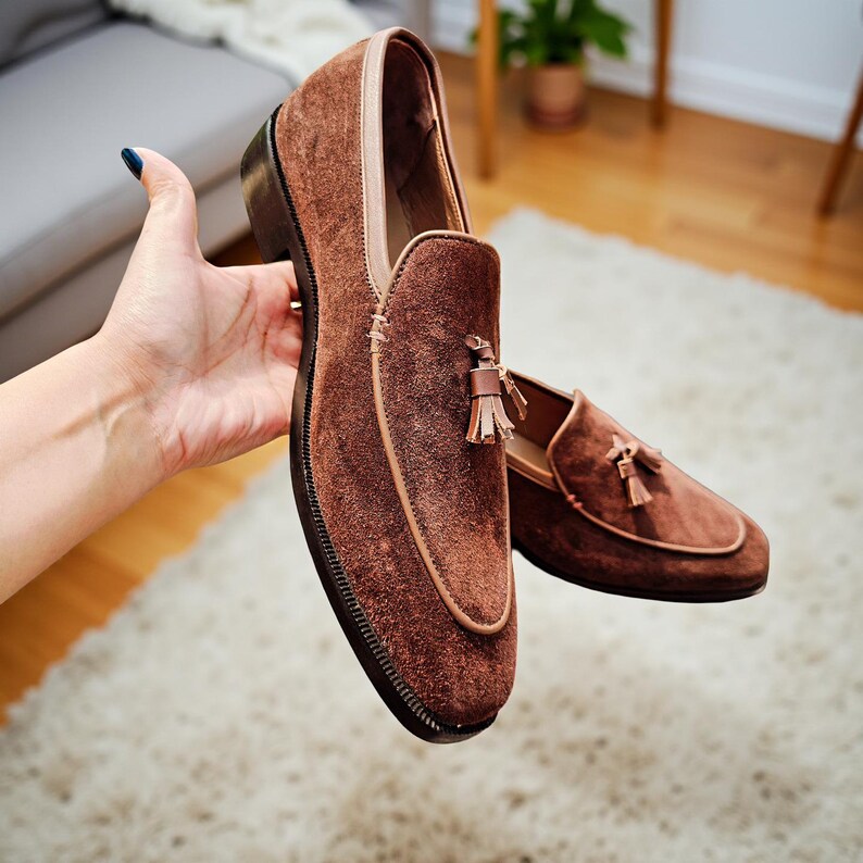 May include: Brown suede loafers with a tassel detail. The shoes have a rounded toe and a leather trim. The sole is dark brown. The shoes are displayed on a white rug.