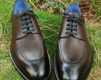 May include: A pair of brown leather dress shoes with black laces and soles. The shoes have a textured upper and a rounded toe design. The interior lining is blue. The shoes are displayed on a bed of green grass.