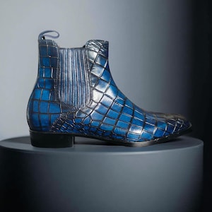 May include: A blue leather Chelsea boot with a crocodile pattern. The boot features a pull tab and elastic side panels. The boot is displayed on a grey cylindrical pedestal. The boot's colour is a vibrant blue.