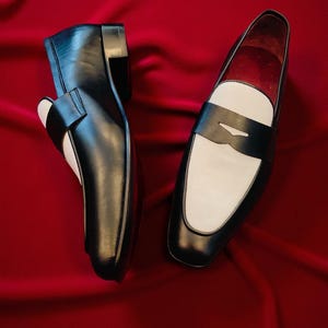 May include: A pair of black and white leather loafers. The shoes feature a classic design with a black upper, a white vamp, and a black strap. The shoes are displayed on a red background.