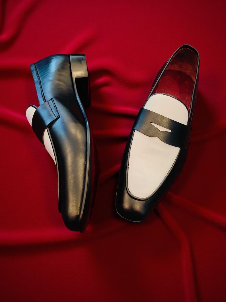 May include: A pair of black and white leather loafers. The shoes feature a classic design with a black upper, a white vamp, and a black strap. The shoes are displayed on a red background.