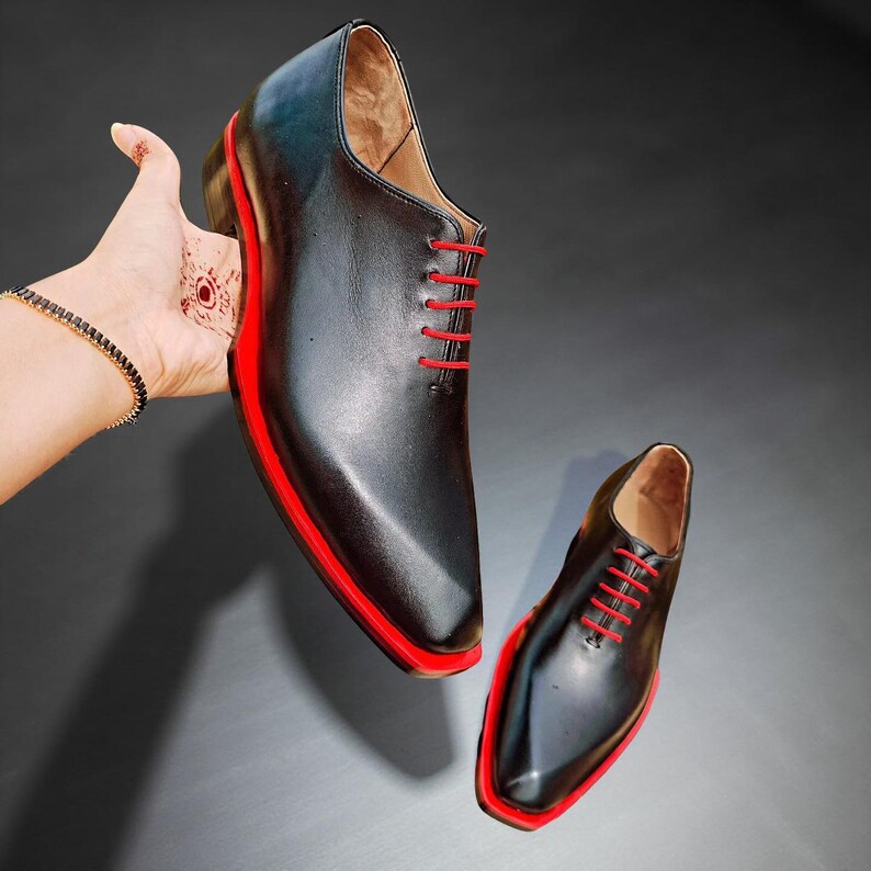 May include: Black leather dress shoes with red soles and red laces. The shoes have a classic design with a modern twist, featuring a sleek silhouette and bold colour accents. A stylish choice for formal occasions.
