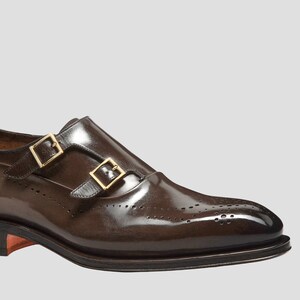 Puede incluir: Primer plano de un zapato marr&oacute;n de cuero con doble hebilla. El zapato presenta dos hebillas doradas y perforaciones decorativas en la puntera. El cuero tiene un acabado brillante.