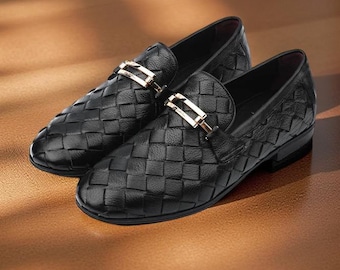 May include: A pair of black leather loafers with a woven texture. Each shoe features a gold-colored buckle detail. The shoes are displayed on a wooden surface, showcasing a classic and elegant design.