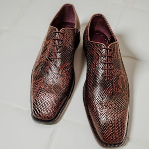 May include: A pair of brown leather dress shoes with a snakeskin pattern. The shoes have a square toe, a lace-up closure, and a dark burgundy interior. The shoes are displayed on a white tiled surface.