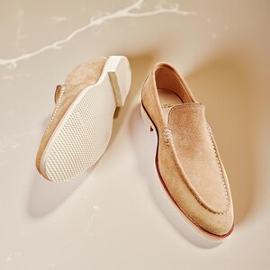 May include: A pair of tan suede loafers. One shoe is upside down, showing the white sole with a textured pattern. The other shoe is upright, displaying the classic loafer design. The shoes are a neutral colour.