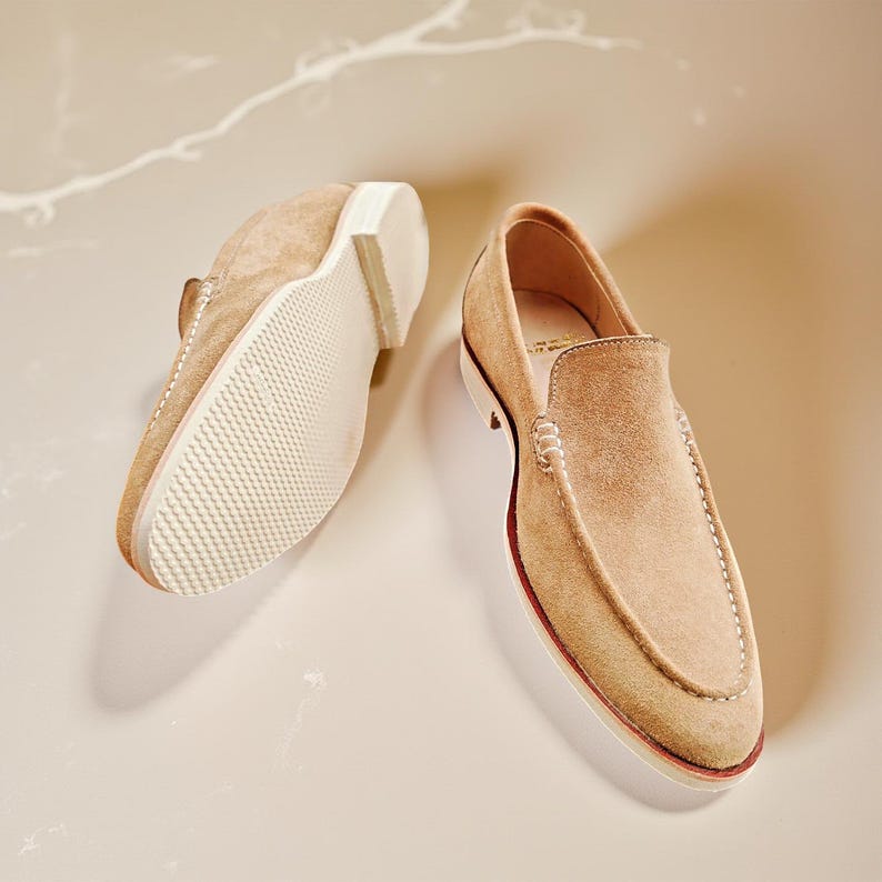 May include: A pair of tan suede loafers. One shoe is upside down, showing the white sole with a textured pattern. The other shoe is upright, displaying the classic loafer design. The shoes are a neutral colour.