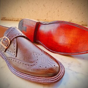 K&ouml;nnte beinhalten: Nahaufnahme eines Paares brauner Lederschuhe. Ein Schuh ist fokussiert und zeigt aufwendige Brogue-Details und einen Schnallenverschluss. Die Sohle des anderen Schuhs ist sichtbar und zeigt eine satte, rotbraune Farbe. Die Schuhe scheinen handgefertigt zu sein.