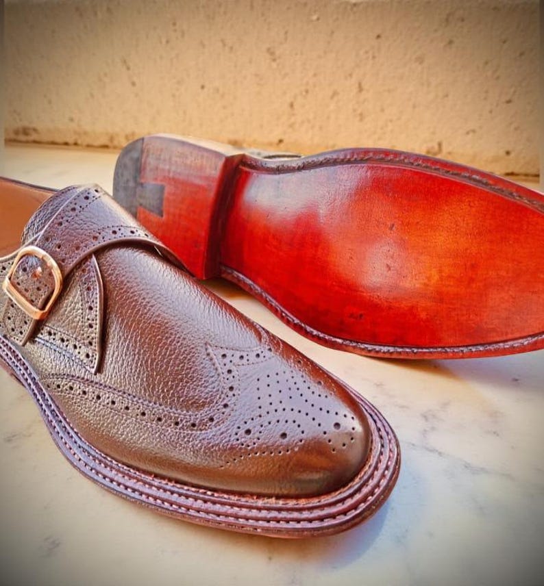 K&ouml;nnte beinhalten: Nahaufnahme eines Paares brauner Lederschuhe. Ein Schuh ist fokussiert und zeigt aufwendige Brogue-Details und einen Schnallenverschluss. Die Sohle des anderen Schuhs ist sichtbar und zeigt eine satte, rotbraune Farbe. Die Schuhe scheinen handgefertigt zu sein.