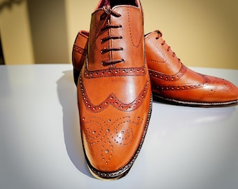 May include: A pair of brown leather Oxford shoes with decorative perforations and wingtip detailing. The shoes have a classic lace-up design and are set against a neutral background. The leather has a polished finish.
