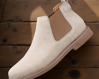 May include: A light beige suede Chelsea boot with a brown rubber sole. The boot has an elastic panel on the side and a pull tab on the back.