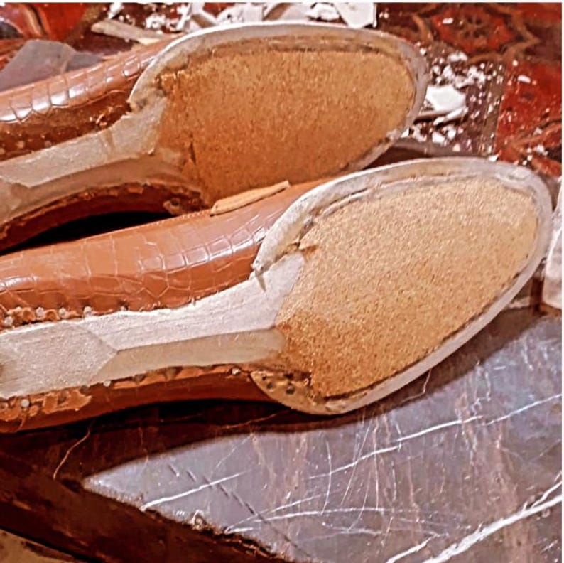 May include: Close-up of the soles of two brown leather shoes. The shoes have a textured, crocodile-like pattern on the upper. The soles are made of a light brown, porous material. The shoes are on a dark, reflective surface.