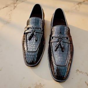 May include: A pair of black leather loafers with a crocodile texture. The shoes have a tassel detail and a silver trim. The loafers are displayed on a light-colored surface.