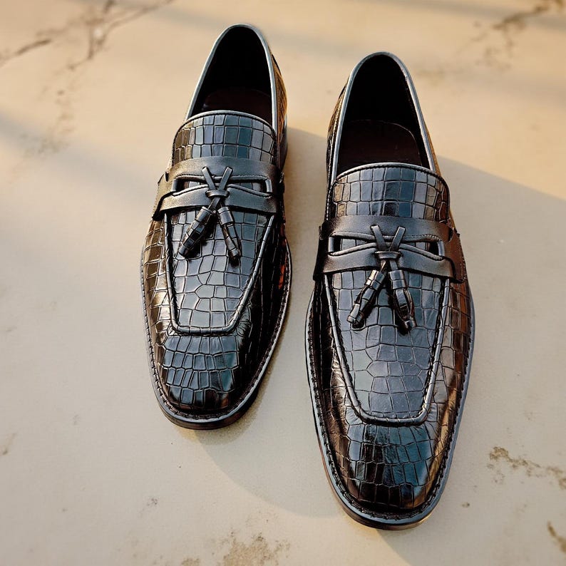 May include: A pair of black leather loafers with a crocodile texture. The shoes have a tassel detail and a silver trim. The loafers are displayed on a light-colored surface.