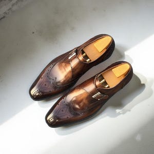 May include: A pair of polished brown leather dress shoes with a monk strap buckle closure. The shoes feature decorative perforations and are displayed with wooden shoe trees inside.