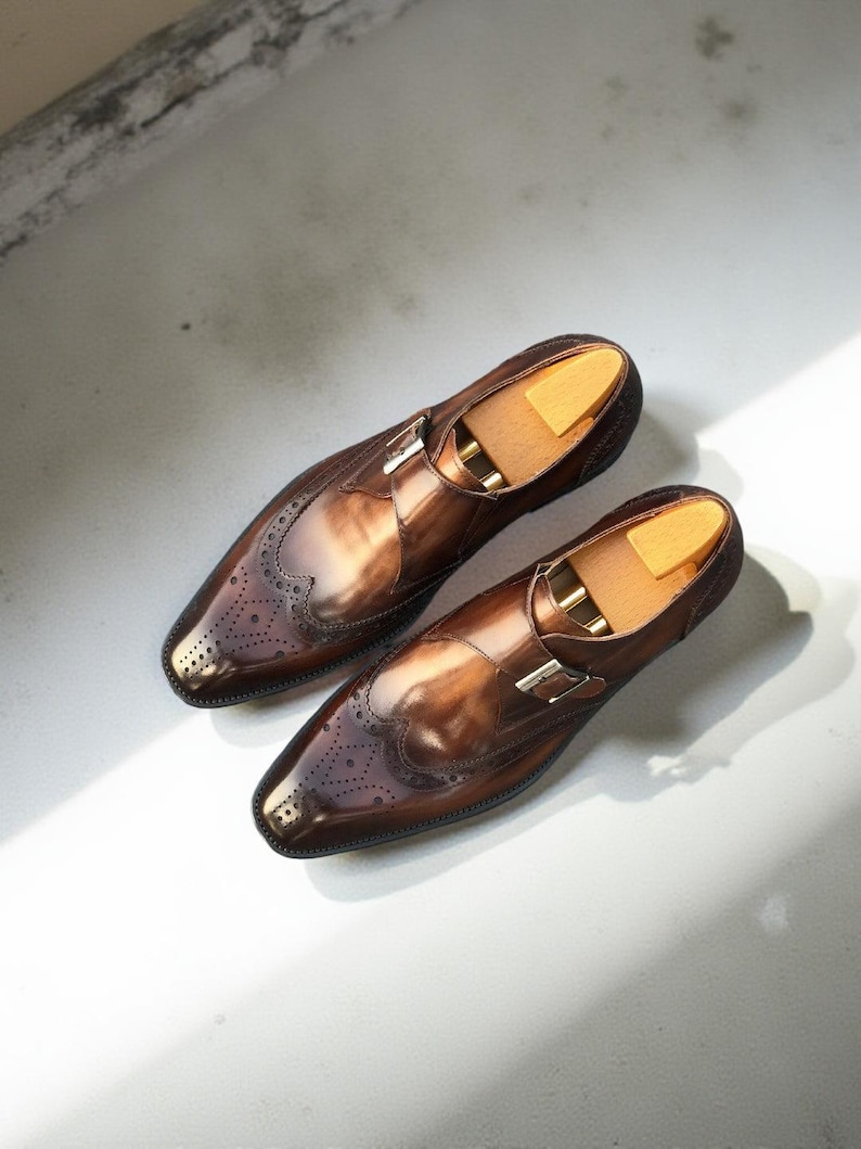 May include: A pair of polished brown leather dress shoes with a monk strap buckle closure. The shoes feature decorative perforations and are displayed with wooden shoe trees inside.