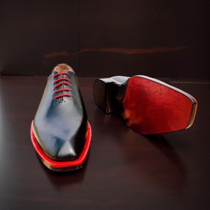 May include: Black dress shoes with red detailing. The shoes feature red laces and a red trim around the sole. One shoe is upright, the other is on its side, showing a red sole.