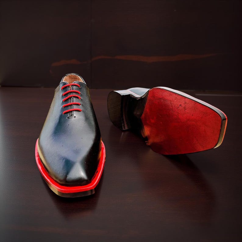 May include: Black dress shoes with red detailing. The shoes feature red laces and a red trim around the sole. One shoe is upright, the other is on its side, showing a red sole.