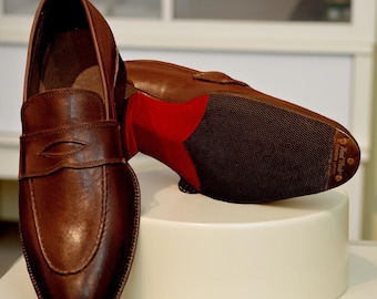 May include: A pair of brown leather loafers with a penny strap design. The soles are dark brown with a red heel and a textured pattern. The shoes are displayed on a white pedestal.