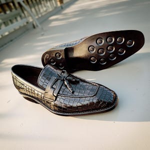 May include: Black leather loafers with a crocodile-like texture. The shoes have a tassel detail and a rounded toe. The sole of one shoe is visible, showing a pattern of circular grips. The shoes are on a light-colored surface.