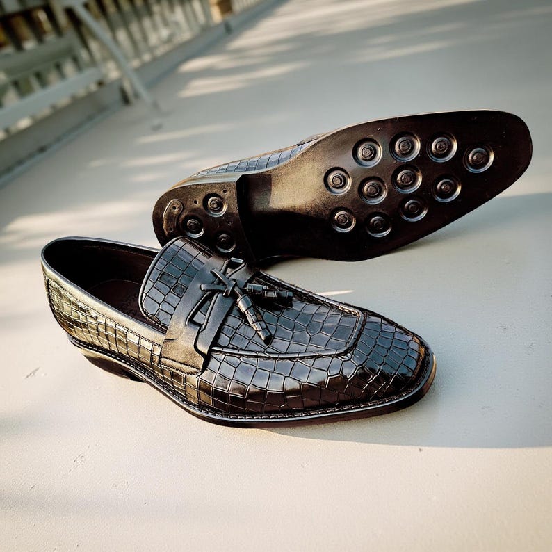 May include: Black leather loafers with a crocodile-like texture. The shoes have a tassel detail and a rounded toe. The sole of one shoe is visible, showing a pattern of circular grips. The shoes are on a light-colored surface.