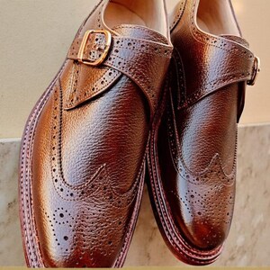 K&ouml;nnte beinhalten: Ein Paar braune Lederschuhe mit Lyralochung und Schnallenverschl&uuml;ssen. Die Schuhe haben eine strukturierte Oberfl&auml;che und ein poliertes Aussehen, mit einer goldfarbenen Schnalle. Die Sohlen sind in einem dunkleren Braunton gehalten.