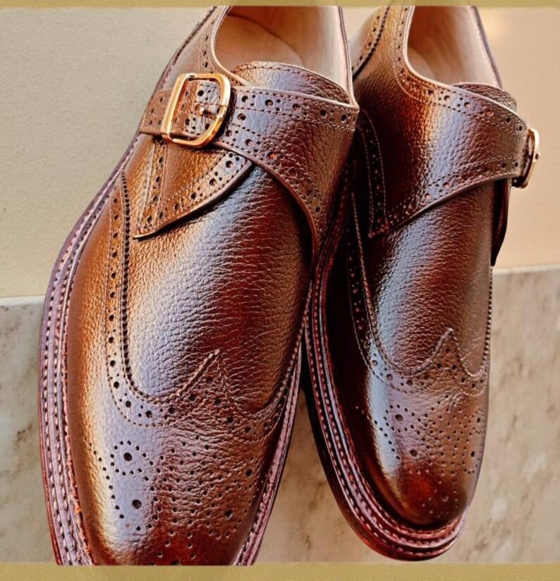 K&ouml;nnte beinhalten: Ein Paar braune Lederschuhe mit Lyralochung und Schnallenverschl&uuml;ssen. Die Schuhe haben eine strukturierte Oberfl&auml;che und ein poliertes Aussehen, mit einer goldfarbenen Schnalle. Die Sohlen sind in einem dunkleren Braunton gehalten.