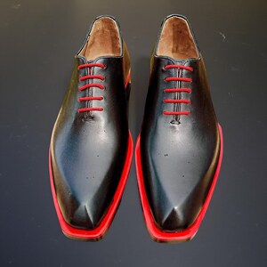 May include: A pair of black leather dress shoes with red soles and red laces. The shoes have a classic Oxford style with a pointed toe. The interior of the shoe is a light brown colour.