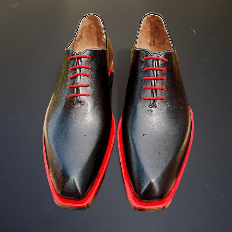 May include: A pair of black leather dress shoes with red soles and red laces. The shoes have a classic Oxford style with a pointed toe. The interior of the shoe is a light brown colour.