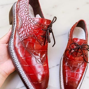 May include: A pair of red leather dress shoes with a textured, crocodile-like pattern. The shoes have a square toe, brown laces, and a white trim around the sole. The shoes are displayed on a white surface.