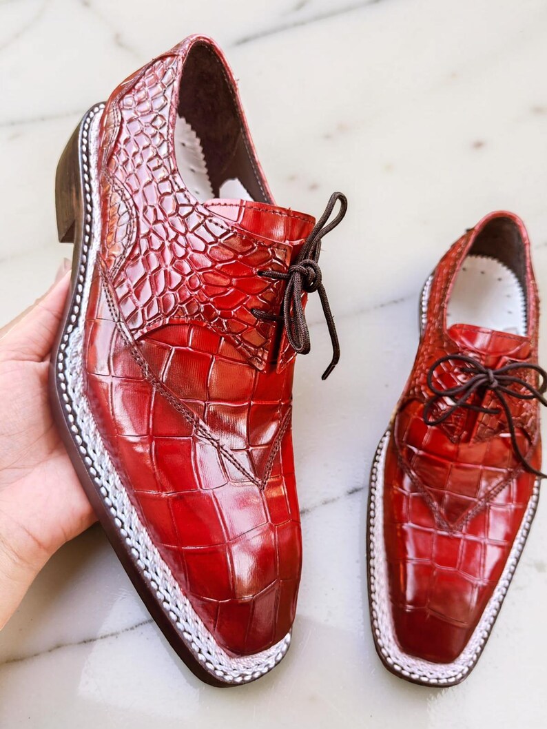 May include: A pair of red leather dress shoes with a textured, crocodile-like pattern. The shoes have a square toe, brown laces, and a white trim around the sole. The shoes are displayed on a white surface.