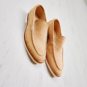 May include: A pair of tan suede loafers with a classic slip-on design. The shoes have a rounded toe, visible stitching, and a contrasting white sole with a red trim. The loafers are displayed on a light-coloured wooden surface.