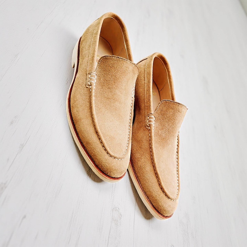 May include: A pair of tan suede loafers with a classic slip-on design. The shoes have a rounded toe, visible stitching, and a contrasting white sole with a red trim. The loafers are displayed on a light-coloured wooden surface.