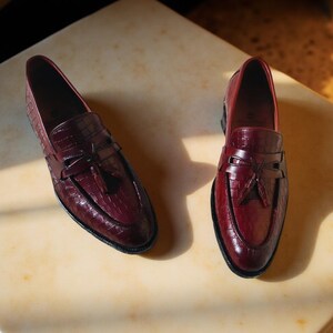 May include: A pair of burgundy leather loafers with a crocodile texture. The shoes have a classic design with a bow detail and a black sole. The loafers are displayed on a light-colored surface.