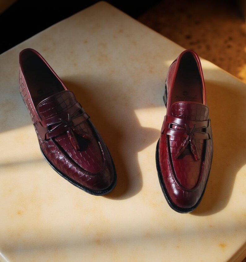 May include: A pair of burgundy leather loafers with a crocodile texture. The shoes have a classic design with a bow detail and a black sole. The loafers are displayed on a light-colored surface.